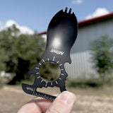 A black metal spork-shaped multi-tool with a circular bottle opener cutout and white measurement markings, held in a hand against a blurred outdoor background of trees and a building.